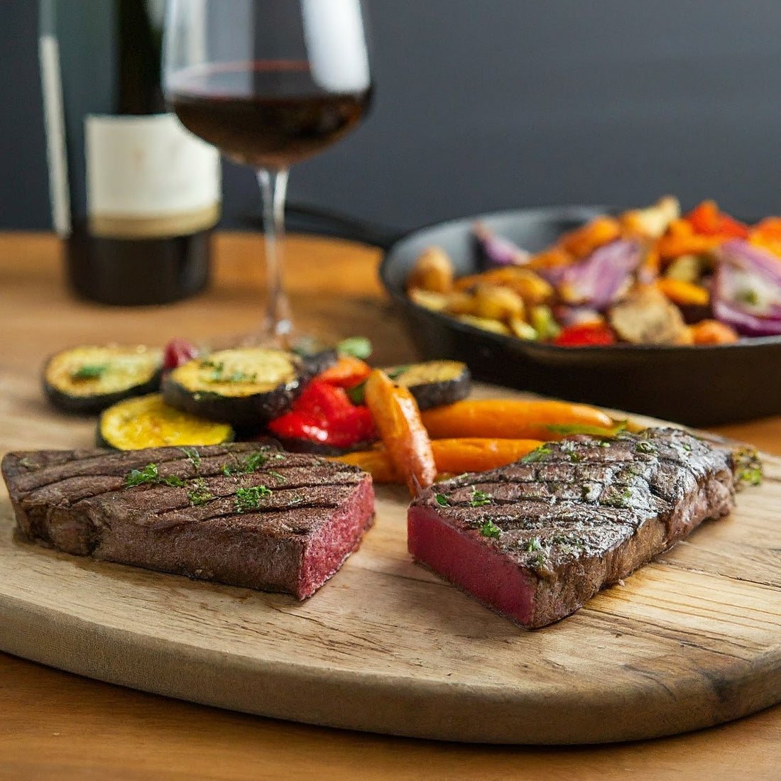 A halved Denver steak with roasted vegetables over a round cutting board with wine and a dish on the side